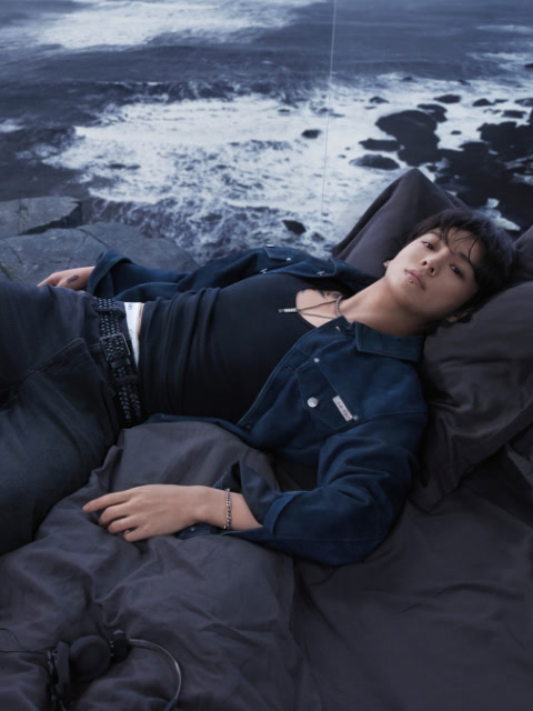 Foto in bianco e nero di una donna che indossa un reggiseno sportivo Calvin Klein e jeans, sdraiata sulla schiena con le gambe piegate sopra la testa, le braccia distese sul pavimento e lo sguardo rivolto alla fotocamera.