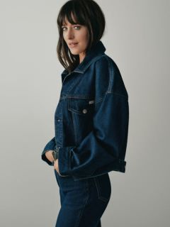 Una persona con lunghi capelli biondi è seduta su una panchina di legno e indossa una giacca di jeans blu, una camicia bianca e jeans larghi. Sullo sfondo si vedono edifici urbani e grattacieli.