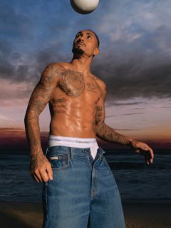 Un uomo con una giacca di jeans fantasia e jeans blu è in piedi su un ponte con ringhiera metallica. Sullo sfondo si vedono un fiume e un altro ponte in una giornata nuvolosa.