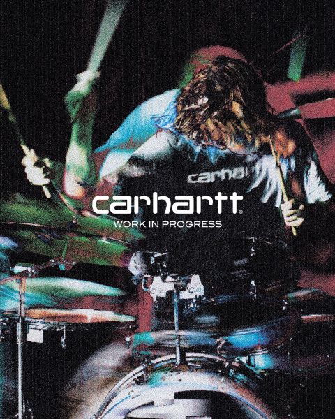 Immagine in bianco e nero di una persona con cappello e giacca accovacciata con le mani sulle ginocchia. Il testo "Carhartt Work In Progress" è ben visibile al centro.
