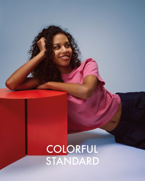 Una persona con lunghi capelli biondi è di profilo su uno sfondo blu. Indossa una maglietta blu scuro, jeans azzurri e scarpe da ginnastica. Il testo "COLORFUL STANDARD" è visualizzato in bianco.