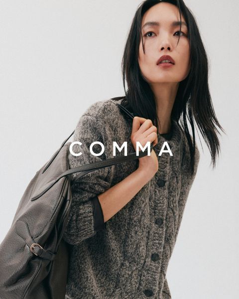 Una donna con un cappotto a quadri e accessori dorati posa con un cane marrone chiaro e bianco in un abito a quadri abbinato. La parola "COMMA" appare in grande carattere bianco su tutta l'immagine.