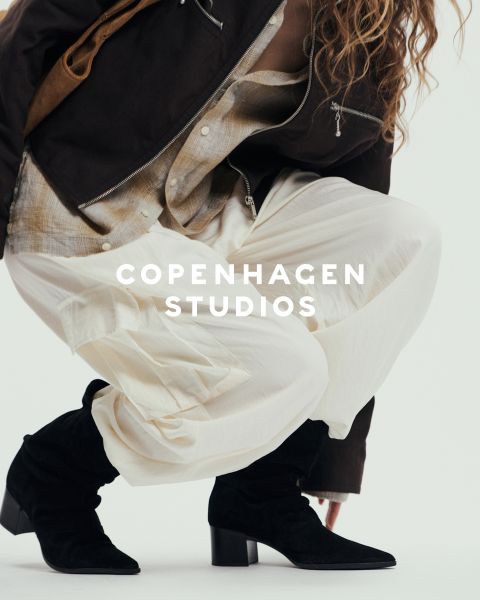 Una persona con stivali stringati neri massicci e pantaloni scuri è in piedi su una superficie bianca. Una parte dell'immagine è oscurata da fili sfocati rosa e beige. Il testo "Copenhagen Studios" appare sopra la foto.