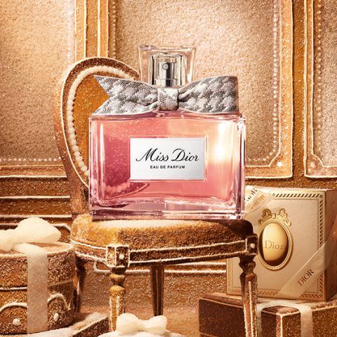 Una bottiglia di vetro trasparente di "Miss Dior Eau de Parfum", riempita con liquido rosa e un fiocco argentato e decorato intorno al collo della bottiglia, su uno sfondo bianco semplice.