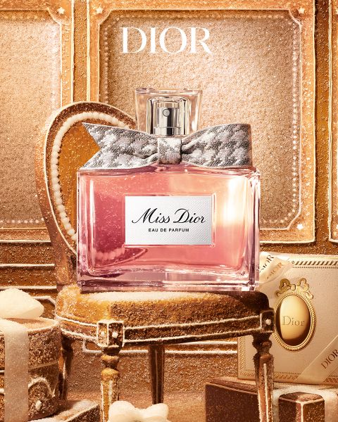Un uomo con tatuaggi e gioielli si appoggia a un tavolo accanto a una bottiglia di profumo "Dior Sauvage Extrait" davanti a uno sfondo esterno accidentato.