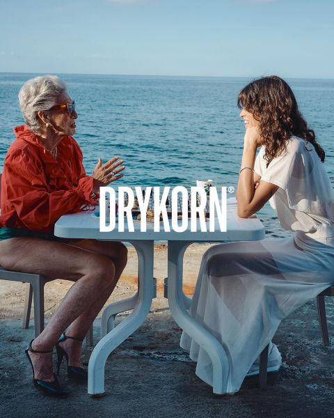 Un'elegante signora anziana con capelli biondo platino e un cappotto di pelliccia nero è seduta su un vecchio divano accanto a un grande cane marrone. La parola "DRYKORN" è visibile nella parte inferiore dell'immagine. L'illuminazione calda crea un'atmosfera lussuosa.