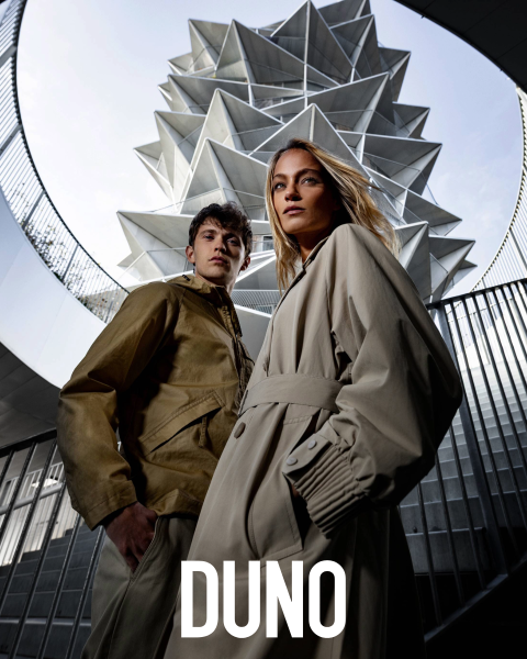 Una persona è di profilo, indossa una giacca giallo chiaro e una gonna bianca, ha la testa reclinata all'indietro e gli occhi chiusi. Si trova in un ambiente esterno con grandi cactus e palme. La parola "DUNO" appare verticalmente sul lato sinistro dell'immagine.