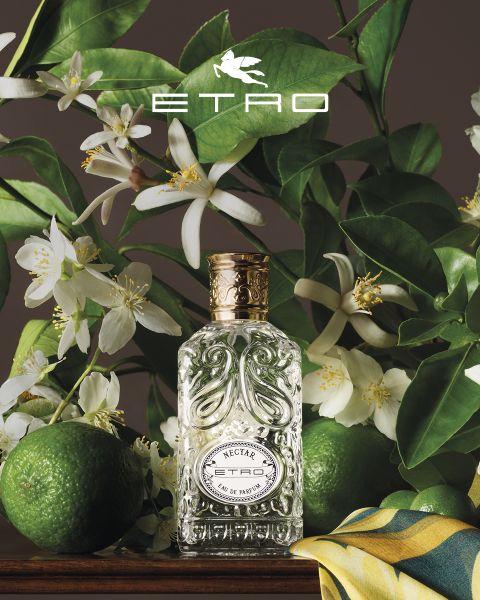 Flacone di profumo con la scritta "ETRO" e un design a pigna, circondato da decorazioni artistiche: una sfera, un uovo con una farfalla, una pietra, una foglia, noci, una mela dorata e una mano scolpita. In alto, il logo ETRO su uno sfondo di cielo blu e mare.