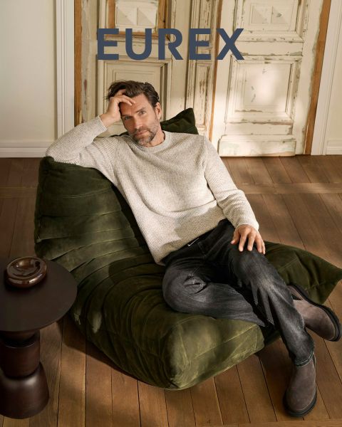 Un uomo è in piedi all'aperto vicino all'erba alta, indossa un maglione beige, un gilet imbottito blu navy e pantaloni marroni e guarda di lato. Il logo "EUREX by BRAX" appare sul lato destro. Il cielo è nuvoloso.