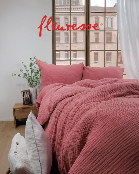 Una camera da letto accogliente con un letto ben fatto con lenzuola bianche con motivo di uccelli. Due cuscini decorativi sono appoggiati alla testiera. La luce del sole filtra attraverso tende trasparenti, proiettando una luce soffusa sul pavimento in legno chiaro.