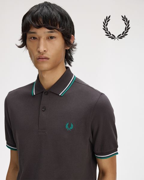 Una persona con lunghi capelli intrecciati guarda pensierosa verso l'alto mentre si sistema una camicia bianca con colletto. La camicia è decorata con una corona d'alloro. L'immagine è in bianco e nero.