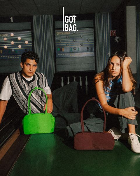 Uomo con occhiali da sole tiene grandi borse grigie davanti a uno sfondo a righe scure. Sotto di lui ci sono diverse borse di diverse dimensioni e colori, tutte esposte in modo ordinato. Il logo in alto a destra è "GOT BAG".
