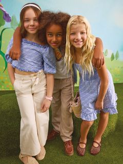 Tre giovani ragazze posano con sicurezza tra le foglie autunnali, indossando abiti metallici lucenti. Due ragazze indossano giacche argentate e la ragazza al centro indossa un top e una gonna di paillettes. Uno sfondo verde acqua contrasta con il fogliame autunnale arancione.