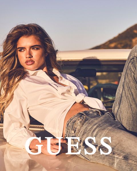 Due donne sono in piedi vicine all'aperto, indossando cappotti strutturati, una in marrone chiaro e una in color crema. Entrambe tengono stretto il colletto e guardano avanti. La parola "GUESS" è scritta in grassetto bianco in basso.