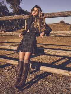 Una donna con lunghi capelli biondi, che indossa un cappotto marrone chiaro e un completo beige a coste, è in piedi davanti a una porta di legno verde e a un muro rosso, decorato con catene e anelli dorati.