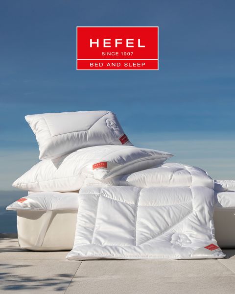 Una donna dorme serenamente su un fianco in un letto moderno con cuscino beige e coperta bianca. Sullo sfondo si vedono un vaso con erba di pampa e una vista sulle montagne al tramonto. In alto campeggia il logo di "Hefel Bed and Sleep".