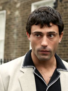 Una persona in abito marrone e con un cappello abbinato è seduta su una panchina nera di fronte a uno sfondo rosso e bianco e guarda di lato con un'espressione pensierosa.