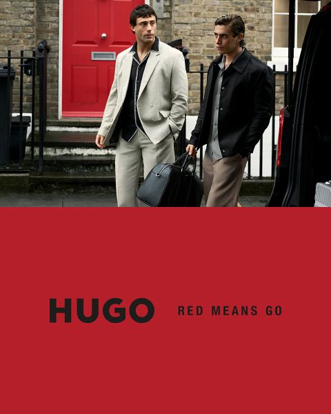 Due modelle in abiti neri posano con sicurezza davanti a uno sfondo chiaro e semplice. Il logo HUGO appare in grassetto bianco nell'angolo inferiore sinistro.