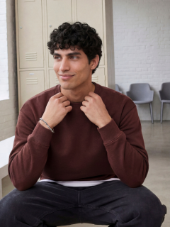 Un giovane uomo con capelli castani corti, che indossa una maglietta color crema con una stampa blu navy sul retro, jeans azzurri e scarpe da ginnastica beige, è seduto sul pavimento e guarda la telecamera sopra la spalla.