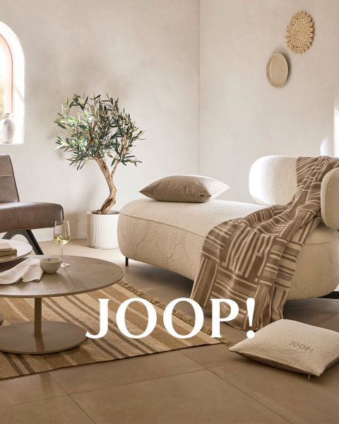Cuscini in colori neutri con motivi e una coperta abbinata sono disposti su una panca minimalista in cemento. Accanto ci sono un grande vaso e una pianta essiccata decorativa. Sulla coperta c'è scritto "JOOP!". Una luce solare soffusa proietta ombre sulla scena.