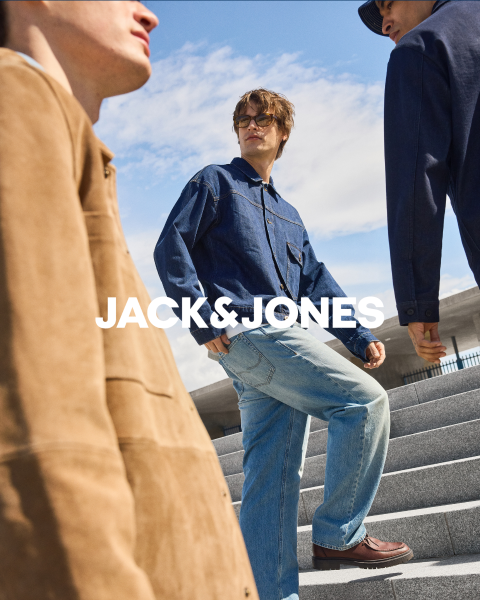 Due uomini in abiti di jeans siedono davanti a uno sfondo semplice. L'uomo davanti si tiene una mano sulla bocca, mentre l'altro è seduto dietro di lui. Al centro c'è il testo "JACK & JONES".