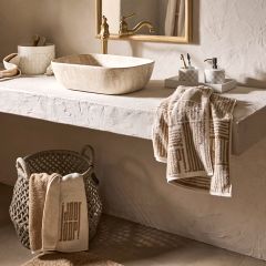 Bagno minimalista con specchio rotondo, rubinetto a parete e lavabo rettangolare. Mensole contengono oggetti decorativi e asciugamani in tonalità naturali. La luce soffusa proietta ombre delicate sulla parete strutturata, creando un'atmosfera serena.