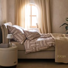 Una camera da letto accogliente con un letto beige con biancheria da letto fantasia. Uno specchio rotondo è appeso al muro. Una luce soffusa illumina una parte del letto. Una piccola pila di libri e oggetti decorativi si trovano nelle vicinanze sul pavimento.