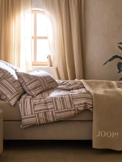 Uno spazio minimalista presenta una panca in pietra beige con due cuscini marroni e beige testurizzati, un plaid a motivi e erbe essiccate. Un cesto intrecciato, un vaso in ceramica rustica e una brocca alta adornano il pavimento sotto una luce naturale soffusa.