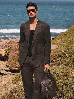 Una persona in un formale abito nero si trova di fronte a una balaustra di pietra con alberi sullo sfondo. Il cielo è parzialmente nuvoloso. La sua camicia è parzialmente sbottonata e ha un'espressione facciale sicura di sé.