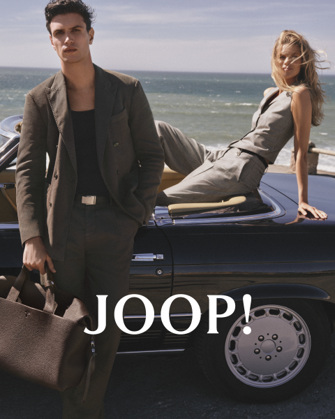 Uomo e donna in abiti estivi alla moda in un caffè. L'uomo indossa una camicia beige e tiene in mano una tazza, mentre la donna con un vestito chiaro tiene un piattino. Entrambi indossano occhiali da sole. "JOOP!" è ben visibile.