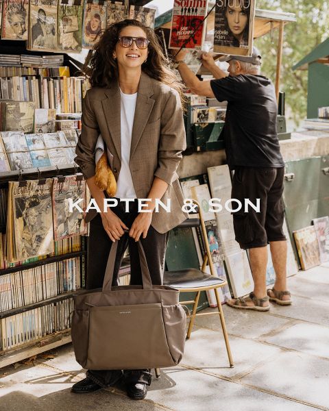 Una donna e un uomo si sorridono all'aperto. La donna indossa un cappello verde e un cappotto beige e tiene in mano una borsa nera. L'uomo indossa una giacca marrone e una borsa color crema. Al centro c'è scritto "Kapten & Son".