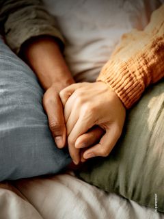 Due persone sdraiate su un letto che si tengono per mano, una con una manica arancione e l'altra con una manica verde, per trasmettere conforto e vicinanza.