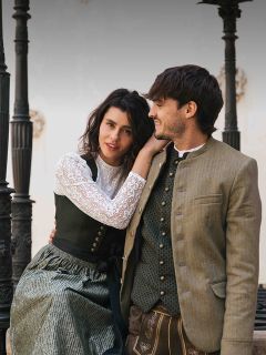 Una donna e un uomo in abiti tradizionali bavaresi sono seduti insieme all'aperto, sorridono e si guardano.