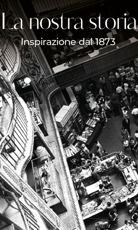 Foto in bianco e nero di un affollato grande magazzino a più piani con persone che fanno acquisti; sono visibili ringhiere decorate e espositori. Il testo sovrapposto in italiano recita: "La nostra storia. Ispirazione dal 1873."