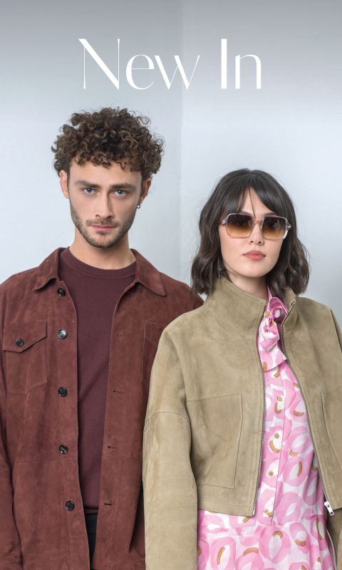 Un uomo con una giacca marrone e una donna con occhiali da sole e una giacca marrone chiaro sopra un abito rosa fantasia sono in piedi uno accanto all'altra davanti a un semplice sfondo chiaro.