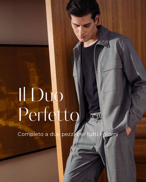 Un uomo in un completo a due pezzi beige chiaro sorride davanti a grandi finestre. Testo: "Il Duo Perfetto. Completo a due pezzi per tutti i giorni."