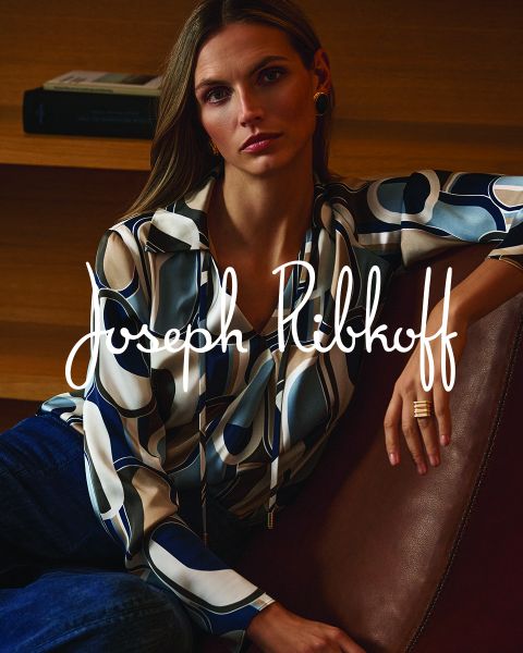 Una donna in una blusa fantasia e jeans è seduta su un divano in pelle; sopra l'immagine è scritto in bianco "Joseph Ribkoff".