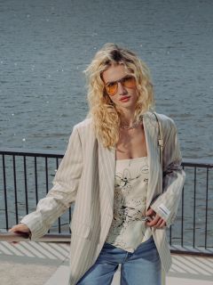 Una donna con capelli biondi ricci indossa occhiali da sole, un blazer a righe, un top floreale, pantaloncini di jeans e scarpe bianche, mentre si trova su una passeggiata con ringhiera.