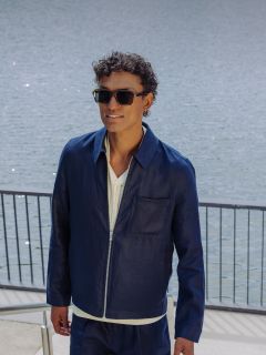 Un uomo con occhiali da sole, giacca blu navy e pantaloncini coordinati sale una scalinata esterna vicino a uno specchio d'acqua, tenendosi a un corrimano e sorridendo.