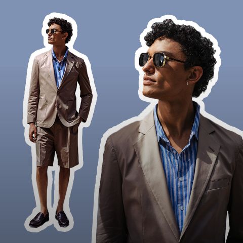 Uomo con occhiali da sole, che indossa un blazer marrone chiaro, una camicia a righe blu e pantaloncini marrone chiaro, in piedi con sicurezza su uno sfondo sfumato marrone.