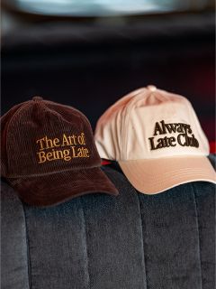 Su un divano grigio ci sono due cappellini da baseball; su quello marrone c'è scritto "The Art of Being Late" e sull'altro beige "Always Late Club".