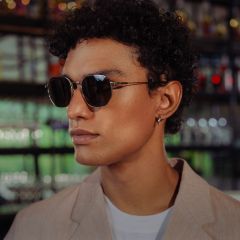 Una persona con i capelli corti e ricci, che indossa occhiali da sole, un blazer beige e una camicia bianca, si trova in un interno con scaffali di vetro sullo sfondo.