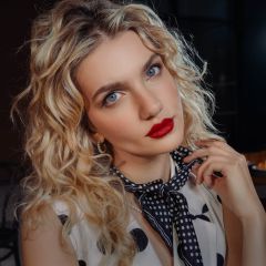 Una donna con capelli biondi ricci, rossetto rosso e una sciarpa a pois guarda la telecamera e tiene la mano dietro il collo.