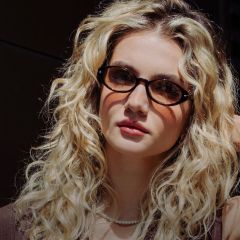 Giovane donna con lunghi capelli biondi ricci, che indossa occhiali tartarugati, una collana di perle e un top marrone, mentre guarda verso la fotocamera.
