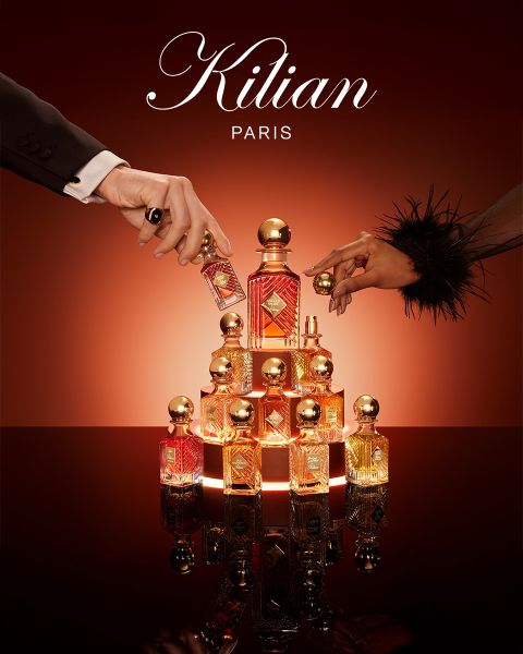 Un flacone del profumo Kilian Paris Angels' Share è accanto a un bicchiere di whisky con ghiaccio e guarnizione, entrambi su una superficie marrone riflettente con illuminazione soffusa. In alto è visibile il logo Kilian Paris.