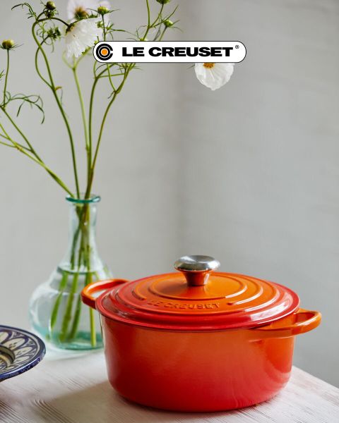 Una scena di cucina con una casseruola rossa, una padella antiaderente, un set di pentole impilabili in acciaio inossidabile, una pentola nera e una ciotola di pomodorini su un bancone di legno chiaro davanti a un paraschizzi di piastrelle bianche. Il logo Le Creuset è visibile.