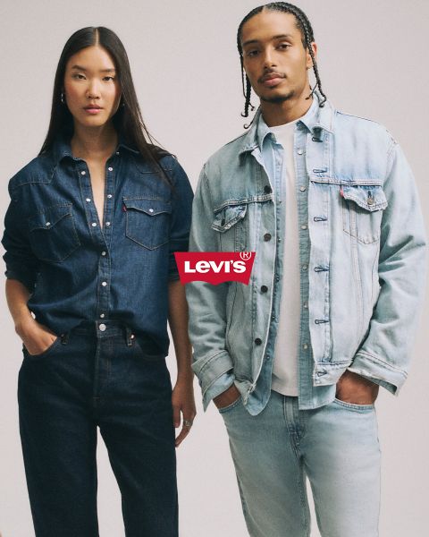 Uomo e donna si abbracciano davanti a uno sfondo patchwork blu, entrambi indossano giacche di jeans e jeans. Sopra di loro spicca chiaramente un logo Levi's rosso.