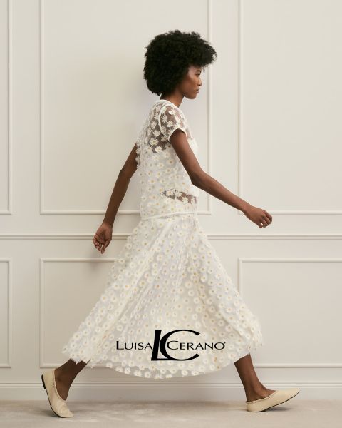 Una donna con lunghi capelli castani indossa un abito a fiori e un capo in pelliccia strutturato color ruggine sulla spalla. Guarda direttamente la fotocamera. In basso al centro si legge il testo "Luisa Cerano".