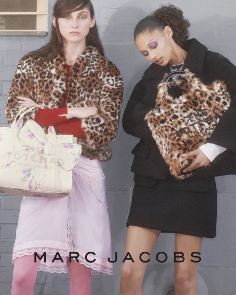 Una donna posa all'aperto con in mano una borsa marrone Marc Jacobs, si appoggia all'indietro con la testa inclinata e indossa una giacca color castagna e jeans blu. Nell'immagine compaiono le scritte "THE TOTE BAG MARC JACOBS" e "MARC JACOBS".