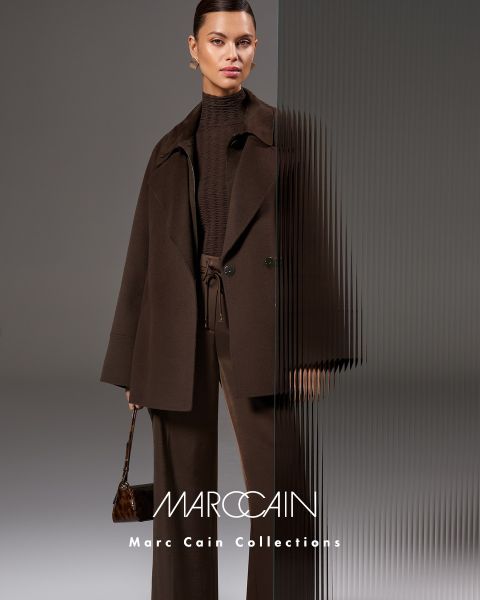 Una donna posa con sicurezza in una giacca di pelle color vinaccia e una gonna con una sciarpa grossa e strutturata color vinaccia. Sullo sfondo si vede un cielo cupo e il marchio "Marc Cain" è visibile in basso.